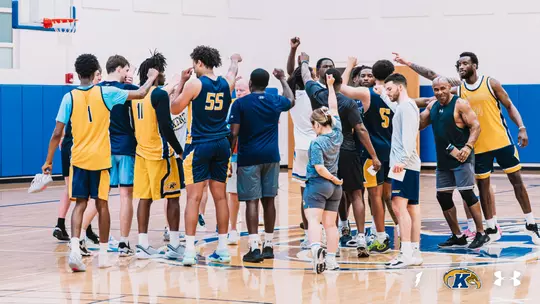 Kent State men's basketball players and staff gather in a tight huddle at center court of the practice gym after practice. Many raise a fist or point upward as they break the huddle. Players wear navy and gold practice jerseys, including numbers 1, 5, 11, 14, and 55, while coaches and staff wear navy, gray, and black team apparel. The court has a Kent State eagle logo at center, with a basket and blue padded walls in the background. Kent State and Under Armour logos appear in the bottom right corner.