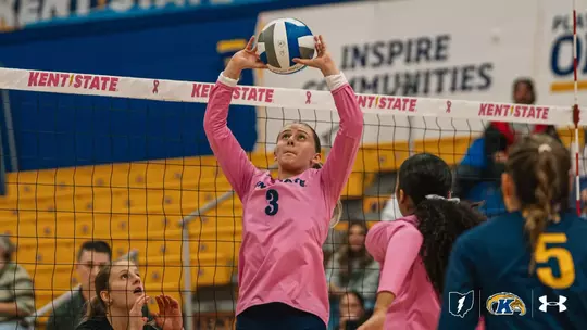 Kent State volleyball player No. 3, wearing a pink long-sleeve jersey, jumps at the net with both hands raised to set the ball overhead during a match in a gym with gold bleachers. Teammates in pink and navy stand nearby, and the net band reads “Kent State” with pink awareness ribbons.