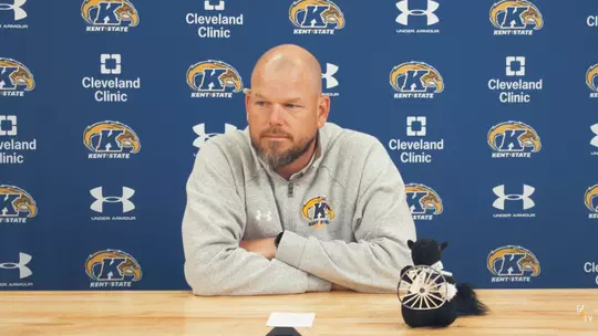A bald man with a beard wearing a gray Kent State pullover seated at a desk during a press conference in front of a blue Kent State backdrop, next to a small black squirrel plush holding a miniature wagon wheel.