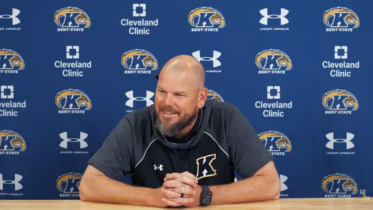 A bald man with a beard wearing a gray Kent State shirt sits with clasped hands during a press conference in front of a blue Kent State backdrop.
