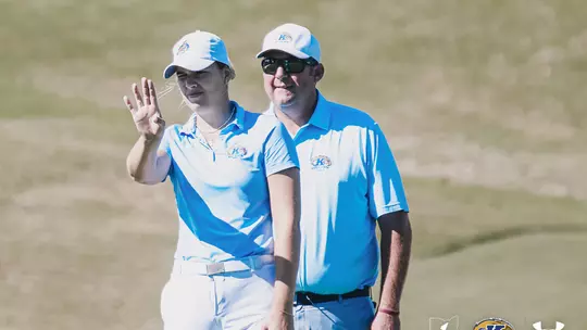 Veronika Kedronová (left) looks toward the green and raises her hand while head coach Casey VanDamme (right) stands beside her, coaching her through a putt; both wear light blue Kent State polos and white caps on the course.