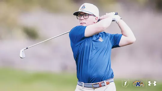 Liam Curtis follows through on an iron shot during a Kent State men's golf practice round. Curtis wears a blue Kent State polo with the Fearless Flash logo, a white cap, glasses, and a white golf glove. Logos for Kent State athletics, the Flashes, and Under Armour appear in the bottom right corner.
