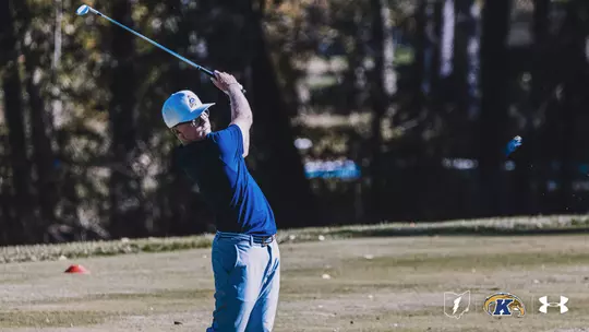 Kent State first-year Liam Curtis follows through on an iron shot during a practice round, wearing a blue polo and white Kent State cap with trees in the background.