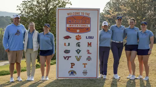 Kent State women's golf team poses on the course next to the Clemson Invitational on Lake Keowee welcome sign, which displays the logos of all participating teams. From left to right: head coach Casey VanDamme, assistant coach Manuela Carbajo Ré, Isabella Goyette, Leon Takagi, Veronika Kedronová, Petra Babicová and Gracie Larsen. The team is wearing Kent State light blue with trees and a hazy sky in the background.