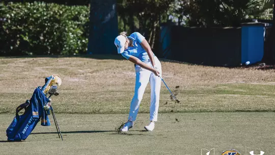 Kent State's Veronika Kedronová swings through an iron shot from the fairway during a sunny round of play, taking a divot as dirt lifts into the air. Her Kent State golf bag stands beside her on the left, with trees and greenery in the background.