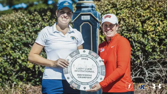 Alt text: Veronika Kedronová of Kent State women's golf poses with the Lady Luck Invitational individual champion trophy alongside a UNLV staff member in a red quarter-zip and white UNLV hat. Kedronová wears a white Kent State polo, blue pants and a navy cap as both smile and hold the large poker-chip-shaped trophy outdoors in Las Vegas.
