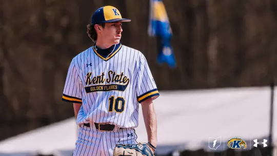 A Kent State Golden Flashes baseball player (Ciaran Caughey) in a striped uniform stands on the field. The uniform features the number 10 and logos for Under Armour and Kent State. A blurred flag and trees are in the background.