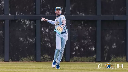 A Kent State University baseball player in a gray road uniform with ‘Flashes' across the chest winds up to throw the ball from the outfield. The player wears a blue cap, a colorful glove, and blue cleats. A green warning track and dark outfield wall are visible in the background. The Kent State ‘K' logo and Under Armour logo appear in the bottom right corner.