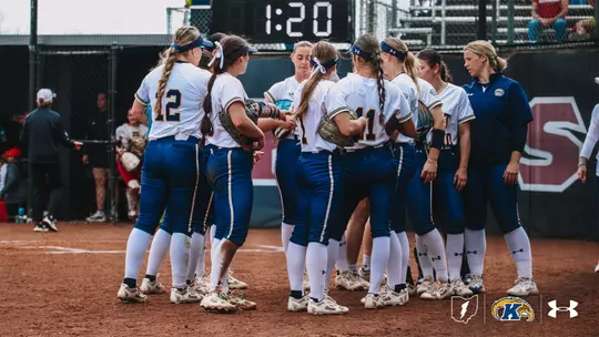 A group of Kent State University softball players in white jerseys and blue pants gather in a huddle on the infield dirt during a game. Several players wear blue headbands and have braided hair. A coach in a navy blue Kent State jacket stands to the right. A digital scoreboard displaying "1:20" is visible in the background. The Kent State "K" logo and Under Armour logo appear in the bottom right corner.