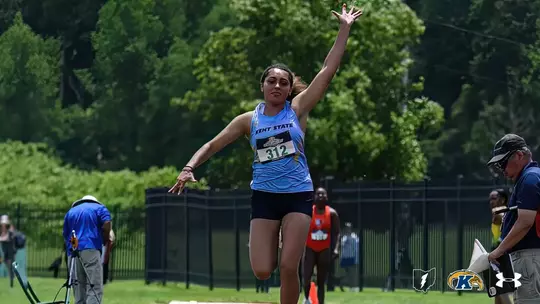 Kent State track and field athlete Lanee Hall, wearing a light blue Kent State uniform with bib number 312, competes in the triple jump, soaring through the air with arms raised and legs extended during an outdoor meet. Officials and spectators are visible in the background, with lush green trees lining the venue.