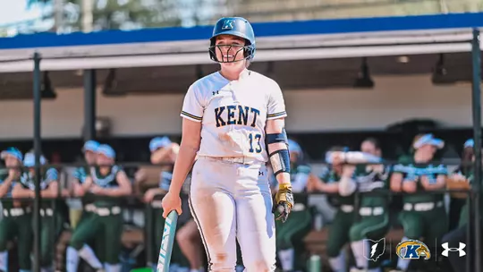 "Kent State softball player Megan Claerhout, wearing jersey number 13, stands at home plate holding a bat during a game. She is dressed in a white Kent State uniform with navy and gold accents, a navy batting helmet with a face guard, gold batting gloves, and a navy arm sleeve. Her uniform shows signs of dirt from play. She is smiling slightly. In the blurred background, the opposing team's dugout is visible, with players in green and white uniforms. The Kent State and Under Armour logos appear in the bottom right corner."