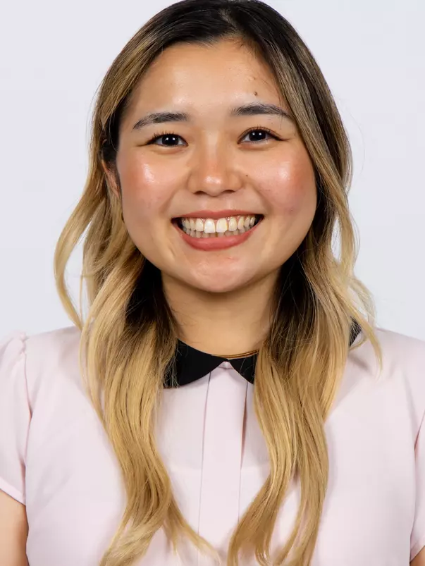 Headshot of Juju Seki smiling broadly, wearing a light pink short-sleeve top with a black collar detail and a gold necklace, with long blonde hair.