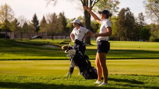 Kent State Dr. Paula Treckel women's golf team first-year Veronika Kedronová holds her hand up to read yardage or wind down the fairway while assistant coach Manuela Carbajo Ré stands behind her, a navy Kent State stand bag between them in the golden late-afternoon light.