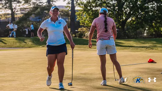 Kent State golfer Aryn Matthews grins and pumps her fist as she walks off the green during the Therese Hession Buckeye Invitational at Ohio State University Golf Club. Matthews is wearing a white Kent State polo, navy skort and blue cap, carrying her putter at her side. An opposing player stands to her right, facing away. Warm late-afternoon sunlight illuminates the green.