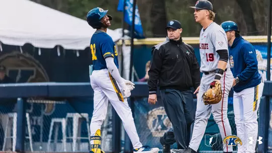 Kent State Golden Flashes baseball player wearing No. 32 in a navy blue jersey and white pants smiles broadly while standing near first base, with an umpire and an opposing player in a white jersey with No. 19 visible in the background, and a Kent State base coach partially visible at right.