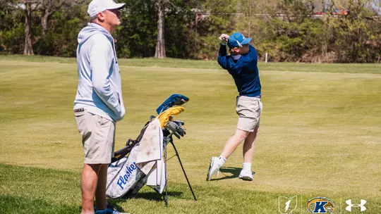 Kent State men's golfer Liam Curtis completes his follow-through on an iron shot in the fairway as head coach Jon Mills watches from behind the team bag. Mills wears a white Kent State cap and hoodie, khaki shorts and blue shoes. Curtis wears a blue Kent State cap, navy long-sleeve pullover, khaki shorts and white golf shoes, with his club finishing high over his left shoulder. The camo-pattern Kent State Flashes stand bag sits between them on the fairway under bright spring sunlight.