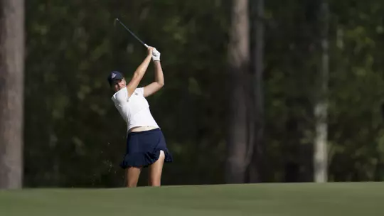 Kent State junior Veronika Kedronová hits an approach shot from the fairway during the second round of the Augusta National Women's Amateur at Champions Retreat Golf Club, wearing a white polo and navy skort with pine trees lining the background.