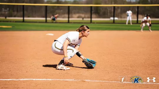 Kent State University softball player Gabby Quinn (#24) crouches low in a ready defensive stance near first base, wearing the team's white and gold Under Armour uniform with a blue fielding glove. Two players are visible in the background in the sunny outfield. The Kent State and Under Armour logos appear in the lower right corner.