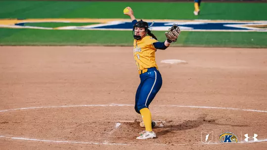 "Kent State softball pitcher Brynn Libler, wearing jersey number 3, winds up to deliver a pitch during a home game. She is dressed in a gold and navy Kent State uniform with gold pants, a black helmet with face guard, and a brown fielding glove. A yellow softball is visible above her head at the top of her wind-up. She is positioned on the pitcher's mound with the Kent State 'K' logo visible on the outfield grass in the background. The Ohio Flash, Kent State Golden Flashes, and Under Armour logos appear in the bottom right corner."