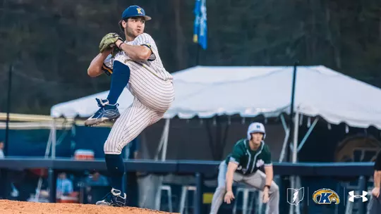 A Kent State Golden Flashes pitcher in a white pinstripe Under Armour jersey, navy socks, and a navy Golden Flashes cap winds up in a high leg kick on the mound, with an opposing baserunner in a green jersey taking a lead in the background.