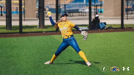 "Kent State softball outfielder Brooke Aberle, wearing jersey number 1, winds up to make a throw from the outfield during a home game. She is dressed in a gold Kent State jersey with navy pants, gold knee-high socks, a navy long-sleeve undershirt, a blue cap, and a tan fielding glove. A yellow softball is visible in her raised throwing hand. She is positioned in a wide stance on the green turf outfield. The stadium seating and fencing are visible in the blurred background. The Ohio Flash, Kent State Golden Flashes, and Under Armour logos appear in the bottom right corner."