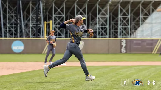 A Kent State Golden Flashes infielder in a gray 'Kent State Golden Flashes' Under Armour jersey and gray pants fires a throw across the diamond, with a teammate visible in the background and the NCAA logo on the outfield wall, during a game at a stadium with exposed steel bleacher framework.