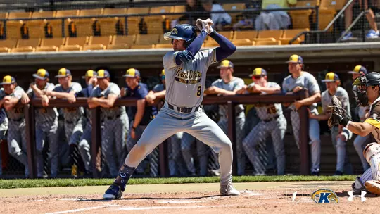Kent State Golden Flashes batter wearing No. 8 stands in his stance at home plate in a gray 'Kent State Golden Flashes' Under Armour jersey and gray pants, with the entire Kent State dugout leaning on the railing watching in the background and an opposing catcher crouched at right.