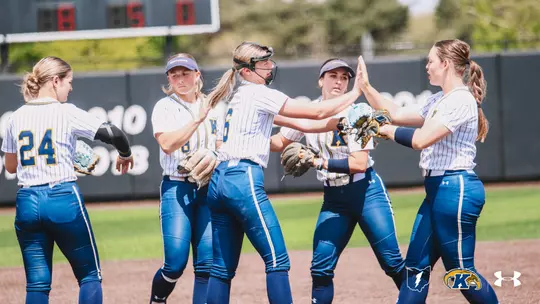 Five Kent State softball players celebrate together on the field during a game, exchanging high-fives in a group. The players are wearing white pinstripe Kent State uniforms with blue pants. Number 24 is visible on the player at far left, and the center player is wearing a catcher's helmet pushed up on her head and holding a fielding glove. A scoreboard showing 8-5 is visible in the background, along with the outfield wall displaying championship years. The Ohio Flash, Kent State Golden Flashes, and Under Armour logos appear in the bottom right corner."