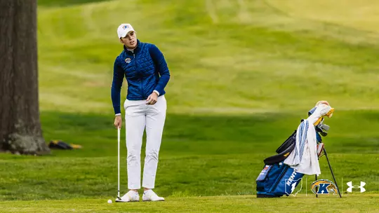 Kent State junior Veronika Kedronová stands over a golf ball on the fairway at Firestone Country Club's Fazio Course during Round 1 of the 2026 MAC Women's Golf Championship, preparing to play a shot. She wears a navy quilted vest, white pants, and a white Kent State Golf cap. A Kent State golf bag with team branding stands to her right, with a tree and open fairway in the background.