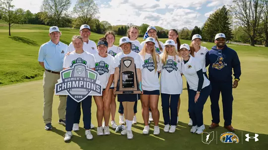 The Kent State Dr. Paula Treckel women's golf team poses on a manicured green at Firestone Country Club's Fazio Course after winning the 2026 Mid-American Conference Women's Golf Championships. Twelve people stand together in two loose rows, smiling at the camera. The player at front center holds the wooden MAC championship trophy, while another player at front left holds a large 2026 MAC Women's Golf Champions shield-shaped sign. Most of the team wears matching white commemorative champions t-shirts and white champions hats, with several in navy Kent State polos. Behind them, a sweeping fairway slopes down to mature trees under a blue sky scattered with white clouds. The Ohio-shaped Golden Flashes logo, Kent State athletic mark, and Under Armour logo appear in the bottom right corner.