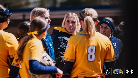 Kent State softball players huddle together during a game against Akron. Several players in gold Kent State jerseys, including number 8, gather closely around teammate Kasey Markusic, who is centered in the frame with a focused expression. A player to the right wears a navy Kent State headband. The dugout is visible in the blurred background. The Ohio Flash, Kent State Golden Flashes, and Under Armour logos appear in the bottom right corner.