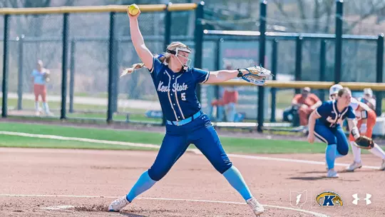 Kent State University softball pitcher Meadow Ferri (#5) is captured mid-windup on the mound, her throwing arm raised high with a yellow softball visible at the top of her delivery. She is wearing the team's navy blue Kent State Under Armour jersey, blue pants, and light blue socks, with a visor and face guard. Her body is in a wide, powerful stride toward the plate. In the background, a teammate can be seen crouched in a ready stance in the infield, along with spectators and a chain-link fence. The Ohio outline, Kent State Golden Flashes, and Under Armour logos appear in the bottom right corner.