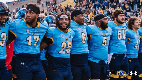 A group of football players wearing blue and yellow jerseys are standing arm in arm on a field. The jerseys display numbers and logos, including the Kent State logo. Spectators are visible in the blurred background, seated in stadium seats.