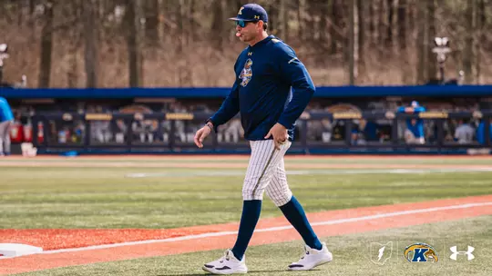 Kent State baseball head coach Jeff Duncan walks along the foul line in a navy Kent State Under Armour pullover, white pinstripe pants, navy socks, a navy gold-brimmed cap, and blue-tinted sunglasses, with the Kent State dugout and baseball wall signage visible in the background.