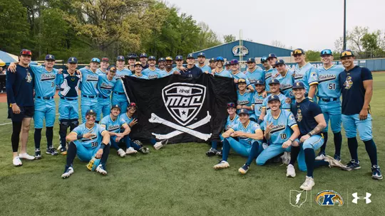 The Kent State baseball team gathers on the field at home for a group photo, holding a large black Mid-American Conference flag with crossed bone, wearing light blue 'Flashes' Under Armour uniforms, with the Kent State baseball facility and championship banner visible on the outfield wall behind them.