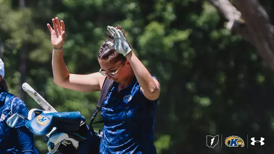 Kent State women's golfer Petra Babicová stands on the course in a navy camo Kent State polo, wearing glasses and a light blue golf glove, with her hands raised above her head as she fixes her hair into a braided style. Her golf bag with Kent State head covers is visible in the foreground, and tall green trees are blurred in the background. The Ohio state logo, Kent State Golden Flashes logo, and Under Armour logo appear in the bottom right corner.