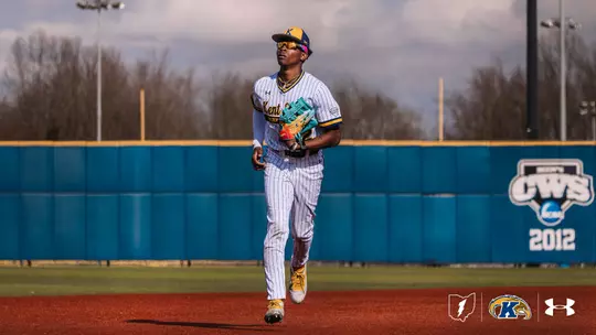 A Kent State Golden Flashes outfielder in a white pinstripe 'Kent State Golden Flashes' Under Armour jersey, white pants, gold cleats, and a navy gold-brimmed cap jogs in from the outfield holding a teal Wilson glove, with the Kent State outfield wall and a 2012 NCAA College World Series banner visible behind him.