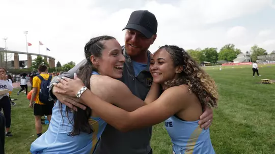 Kent State track and field assistant coach Michael Schober embraces two student-athletes in a group hug on the infield following a competition. A Kent State track and field coach embraces two athletes in a group hug on the infield following a competition. The coach, wearing a dark gray Under Armour quarter-zip and black cap, smiles broadly while hugging the two athletes, who are dressed in light blue and gold Kent State uniforms. Both athletes are smiling and laughing. A track facility with flags, a curved architectural structure, and green trees is visible in the background, with other competitors and spectators milling about.