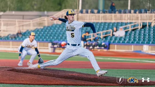Kent State Golden Flashes pitcher wearing No. 5 extends fully through his delivery on the mound in a white pinstripe 'Kent State Golden Flashes' Under Armour jersey, navy sleeves, and a navy gold-brimmed cap, with a teal glove, a teammate set at an infield position behind him, and a partially filled stadium with teal seats visible in the background.