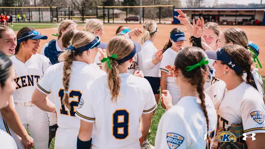 A wide action photo of the Kent State softball team gathered in an energetic team huddle on an outdoor softball field on a sunny day. The players are wearing white Kent State home uniforms with navy and gold lettering, navy and gold visors, and green ribbon hair ties and accessories throughout the group. Several players are visible from behind, including jersey numbers 8 and 12. Players on the right side of the huddle have their hands raised in celebration, with one player making a hand gesture above the group. A catcher's glove is visible on one player at the left. Trees, a red pickup truck, and a dugout structure are visible in the blurred background. The MAC Conference logo appears on the jerseys. The MAC Conference logo, Ohio state outline with lightning bolt, Kent State Golden Flashes logo, and Under Armour logo are displayed in the bottom right corner of the image.