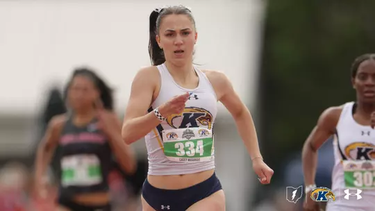 Casey Musgrave of Kent State drives through a sprint event, wearing a white Kent State Under Armour tank top and navy shorts with bib No. 334. A teammate wearing bib No. 303 and a competitor in a dark uniform are visible in the background. The Kent State Athletics logo and Under Armour wordmark appear in the lower right corner.