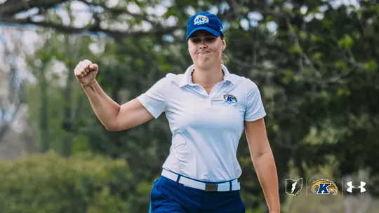 Veronika Kedronová pumps her fist in celebration on the golf course. She wears a white Kent State golf polo, blue Kent State cap and blue pants, with mature trees in soft focus behind her. The Kent State Athletics Ohio mark, primary athletic logo and Under Armour logo are arranged in the bottom-right corner.