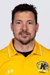 Headshot of Dave Bucar smiling, wearing a gold Kent State Football Under Armour polo shirt with the Golden Flashes logo and white collar trim, with short dark hair and light stubble.