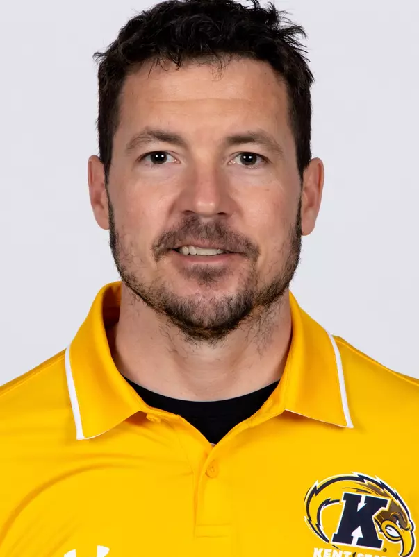 Headshot of Dave Bucar smiling, wearing a gold Kent State Football Under Armour polo shirt with the Golden Flashes logo and white collar trim, with short dark hair and light stubble.