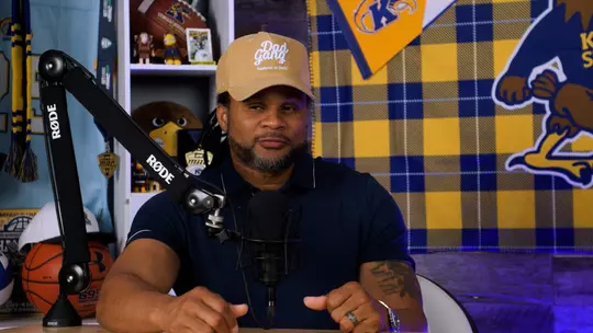 Josh Cribbs sits at table with hat that says Dad Gang. Part of a pennant with the Kent State K logo is in the background