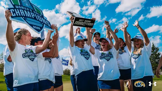 The Kent State women's golf team celebrates on the course, raising their arms and cheering while holding the 2026 MAC Women's Golf Champions trophy and a "Get Some MACtion" sign. The team wears white MAC Women's Golf Champions T-shirts and blue skirts. The Kent State and Under Armour logos appear in the bottom right corner.