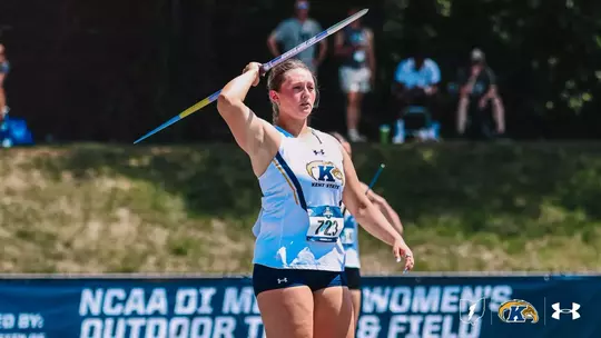 Kent State javelin thrower Kristen Jay winds up for her throw at the NCAA Division I Men's and Women's Outdoor Track and Field Championships. Wearing bib No. 723 and a white Kent State Under Armour uniform, she holds the blue-and-yellow javelin above and behind her head, eyes focused forward. Spectators and a grassy embankment are visible in the background.