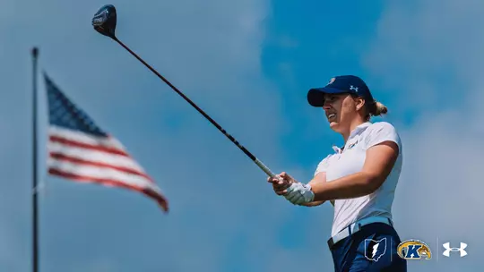 A Kent State women's golfer holds her follow-through and watches her drive against a bright blue sky, wearing a white Kent State polo, navy pants, a navy Under Armour cap, and a golf glove, with an American flag waving in the background and the Kent State lightning bolt, Golden Flashes, and Under Armour logos in the lower right corner.