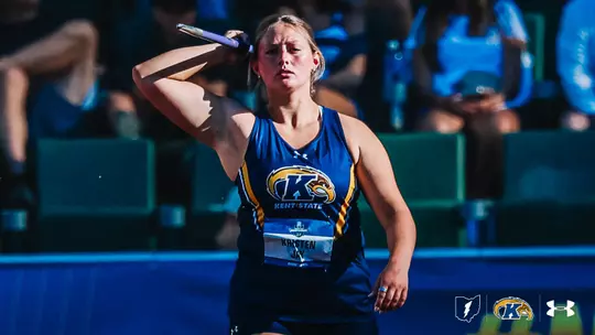 Kent State javelin thrower Kristen Jay winds up for a throw at the NCAA Division I Outdoor Track and Field Championships, holding the javelin cocked behind her right shoulder as she prepares to release. She wears a navy blue Kent State Under Armour uniform with gold accents and the Golden Flashes logo on the chest, along with a competition bib reading "KRISTEN JAY." Blurred spectators and teal stadium seating fill the background. The Kent State Athletics logo, Golden Flashes wordmark, and Under Armour logo appear in the bottom-right corner.