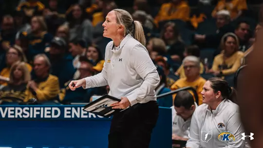 Kent State women's basketball head coach Fran Recchia stands on the sideline holding a clipboard, looking intently toward the court during a game.Kent State women's basketball head coach Fran Recchia stands on the sideline holding a clipboard, looking intently toward the court during a game. She wears a white Under Armour pullover with "Kent Basketball" lettering in gold and black pants, her blonde hair pulled back in a ponytail. A staff member in a matching white Kent State Under Armour jacket is seated behind her. The arena is filled with fans wearing gold and navy. A blue LED courtside advertising board reading "VERSIFIED ENER…" is visible in the background. The Kent State Golden Flashes logo, Ohio state outline, and Under Armour wordmark appear in the lower right corner.